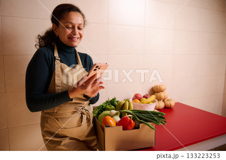 Smiling Woman With Apron Checks Her Phone Beside Fresh Produce Box On Red Counter 133223253