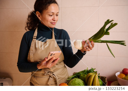 Woman In Apron Checks Her Phone While Holding Leek And Onion In Kitchen Setting 133223254