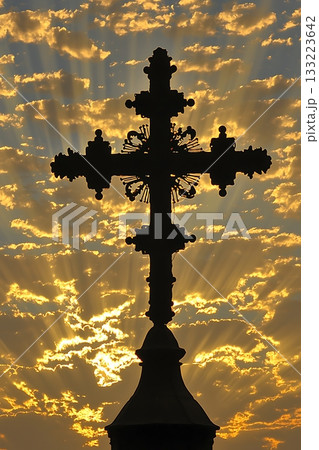 Cross is silhouetted against a backdrop of clouds and a setting sun 133223642