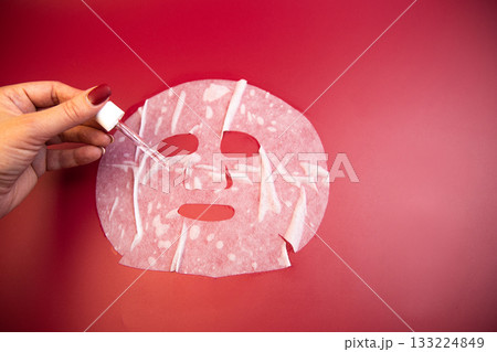 Hand with dropper applies serum to facial mask on bright red background, emphasizing skincare routine and beauty treatment for rejuvenating and nourishing skin 133224849