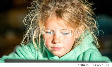 Young girl focused on laptop screen with bright sunlight illuminating face 133225075