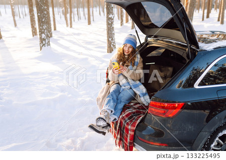 Happy Woman sitting in trunk, using phone. Snowy weather. Concept of adventure, travel by car. Happy Woman sitting in trunk, using phone. Snowy weather. Concept of adventure, travel by car. 133225495