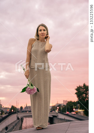 Woman in a knitted beige dress holds a pink hydrangea against a pastel sunset sky over the city 133225596