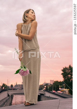 Woman in a knitted beige dress holds a pink hydrangea against a pastel sunset sky over the city 133225597