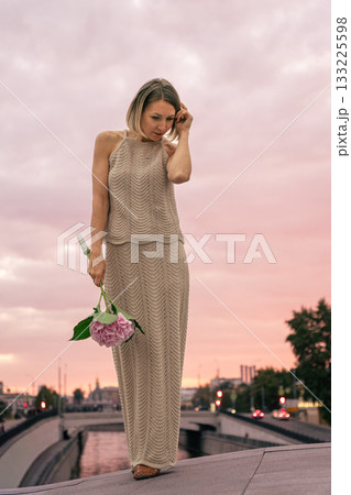 Woman in a knitted beige dress holds a pink hydrangea against a pastel sunset sky over the city Woman in a knitted beige dress holds a pink hydrangea against a pastel sunset sky over the city 133225598