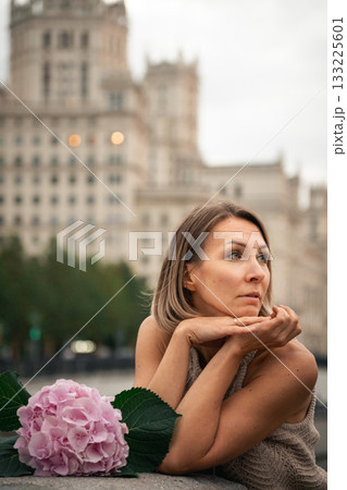 Woman in a knitted dress leans on a stone railing with a pink hydrangea flower in the foreground and a grand city building in the background Woman in a knitted dress leans on a stone railing with a pink hydrangea flower in the foreground and a grand city building in the background 133225601
