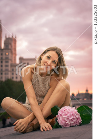 Woman in a beige knitted dress sits thoughtfully on a stone surface at sunset with a pink hydrangea beside her 133225603