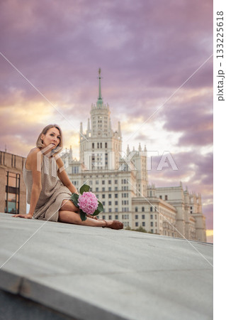 Woman in a beige knitted dress sits gracefully on a stone surface, holding a pink hydrangea against a pastel sunset sky Woman in a beige knitted dress sits gracefully on a stone surface, holding a pink hydrangea against a pastel sunset sky 133225618