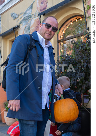 Father kneels beside his child holding a pumpkin amid outdoor seasonal decor. Bright expressions and urban storefront details combine to produce warm family-travel imagery with editorial potential 133225936