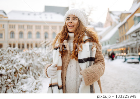 Young happy woman in a winter coat and scarf walks along a snowy city street. Concept of style. 133225949