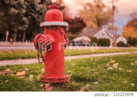 Red fire hydrant on a quiet suburban street in the USA during autumn, surrounded by green grass and fallen leaves at sunset. Red fire hydrant on a quiet suburban street in the USA during autumn, surrounded by green grass and fallen leaves at sunset. 133225976