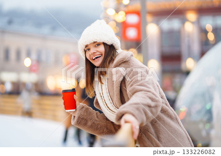 Happy woman with disposable paper coffee cup in winter over outdoor ice skating rink on background. Happy woman with disposable paper coffee cup in winter over outdoor ice skating rink on background. 133226082