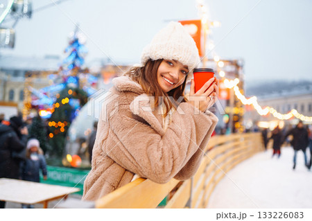 Happy woman with disposable paper coffee cup in winter over outdoor ice skating rink on background. 133226083