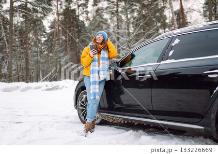 Young woman with phone near her car in a winter forest. Sending messages, communicating with friends 133226669
