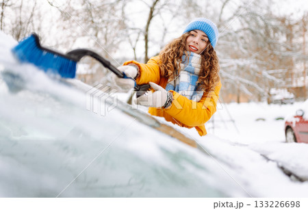 Woman cleaning snow off car during winter snowfall with brush. Scraping ice. Winter cleaning car. Woman cleaning snow off car during winter snowfall with brush. Scraping ice. Winter cleaning car. 133226698