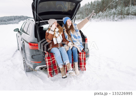 Two young women near a car on a snowy road. Traveling in Winter. 133226995