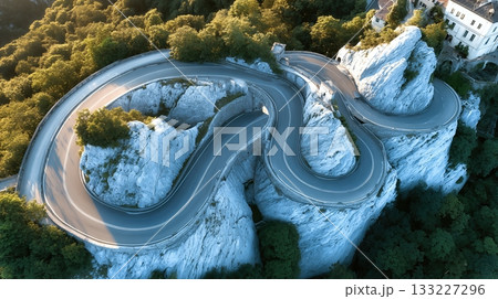 Winding Asphalt Road Curves Through Mountains With Lush Greenery Surrounding Rocky Cliffs Winding Asphalt Road Curves Through Mountains With Lush Greenery Surrounding Rocky Cliffs 133227296