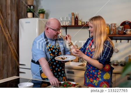 White couple sampling homemade dish together, man offers spoon while woman evaluates flavor, wooden shelves, warm kitchen lighting, thoughtful expressions and casual White couple sampling homemade dish together, man offers spoon while woman evaluates flavor, wooden shelves, warm kitchen lighting, thoughtful expressions and casual 133227765