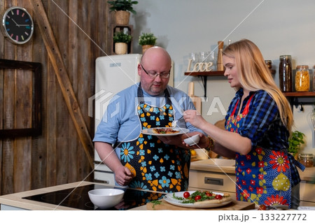 White couple cooking together in kitchen, man tasting dressing while woman serves plate, colorful aprons, wooden countertop, fresh vegetables, warm lighting, candid White couple cooking together in kitchen, man tasting dressing while woman serves plate, colorful aprons, wooden countertop, fresh vegetables, warm lighting, candid 133227772
