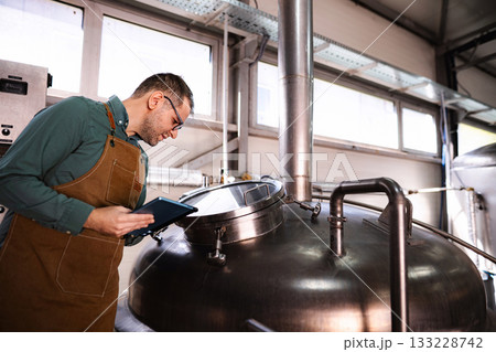 Man wearing apron checking beer fermentation on tablet in microbrewery Man wearing apron checking beer fermentation on tablet in microbrewery 133228742