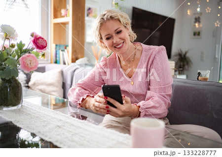 Woman smiling while using phone in living room Woman smiling while using phone in living room 133228777