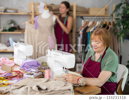 Mature dressmaker sewing clothes against the background of assistant and mannequin 133231318