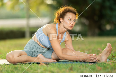 Woman practicing hatha yoga in garden, stretching in an asymmetrical seated asana Parivritta Janu Sirsasana 133231888