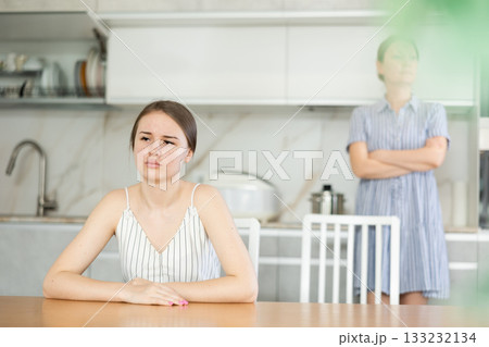 Offended young girl sitting at the kitchen table, angry mother standing behind Offended young girl sitting at the kitchen table, angry mother standing behind 133232134