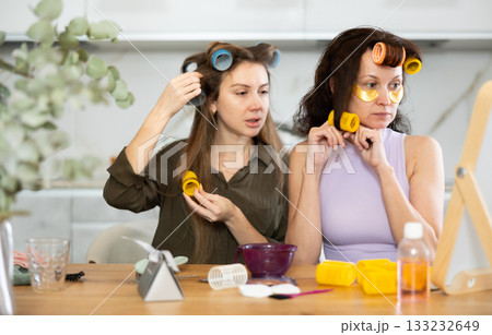 Two adult besties applying hair curlers in front of tabletop mirror 133232649