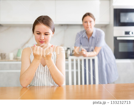Offended young girl sitting at the kitchen table, mother standing behind 133232713