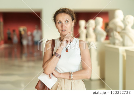 Thoughtful woman visiting museum exhibition of statues with information booklet in hand Thoughtful woman visiting museum exhibition of statues with information booklet in hand 133232728