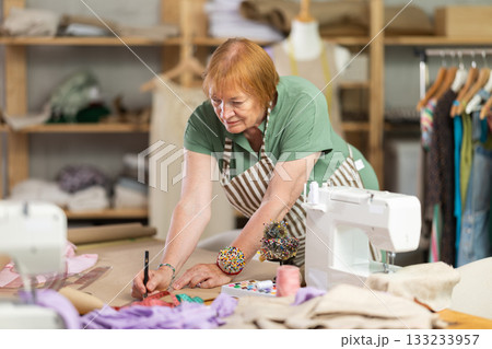 Dressmaker woman works in a sewing workshop and creates patterns of clothes on paper. Tailor with a measuring tape creates fashion design 133233957