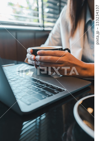 Close-up of a person holding a coffee cup while using a laptop in a contemporary office environment. Close-up of a person holding a coffee cup while using a laptop in a contemporary office environment. 133234337