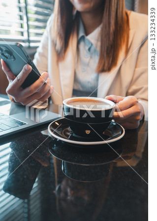 Professional woman in office attire holding a smartphone with a cup of coffee and laptop on a glossy table. 133234339