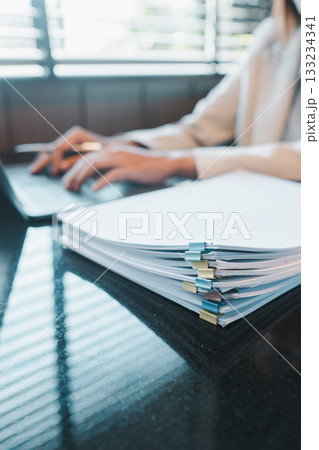 Close-up of a professional working on a laptop with a stack of documents in a modern office environment. Close-up of a professional working on a laptop with a stack of documents in a modern office environment. 133234341