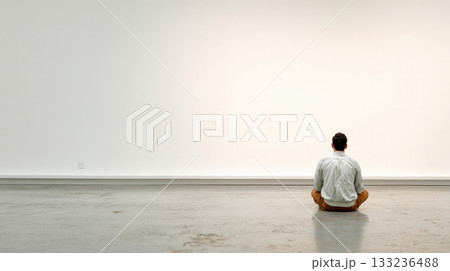 Man sitting alone on floor facing empty white wall, contemplative and calm in modern interior 133236488