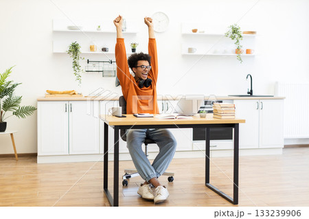 A young man with glasses celebrates a win while working on his laptop at home. A young man with glasses celebrates a win while working on his laptop at home. 133239906