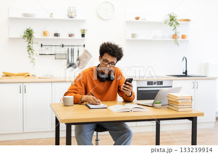 A young man with glasses is working from home in his kitchen, using his phone and taking notes. A young man with glasses is working from home in his kitchen, using his phone and taking notes. 133239914