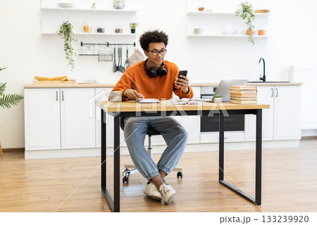 A young man with glasses is working on his laptop and phone at a table in his kitchen. A young man with glasses is working on his laptop and phone at a table in his kitchen. 133239920
