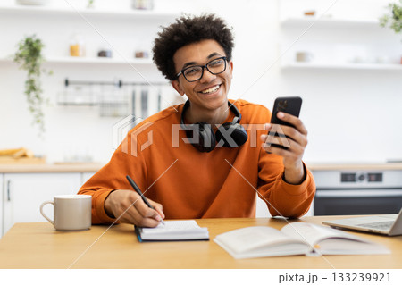 A cheerful young man with glasses smiles while holding a phone and writing in a notebook at a table. A cheerful young man with glasses smiles while holding a phone and writing in a notebook at a table. 133239921