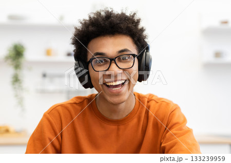A cheerful young Black man wearing glasses and headphones smiles broadly, looking directly at the camera. 133239959