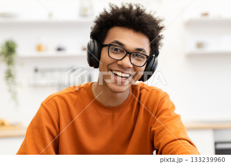 A young man with glasses and headphones smiles brightly at the camera, wearing an orange sweater. 133239960