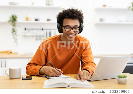 A young man wearing headphones and glasses smiles while studying with a laptop and taking notes in a notebook at home. A young man wearing headphones and glasses smiles while studying with a laptop and taking notes in a notebook at home. 133239963