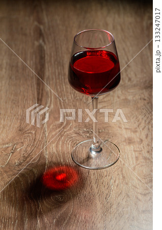 Rich red liquid fills elegant glass casting a playful shadow on warm wooden surface 133247017