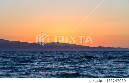 Majestic sunset over tranquil ocean waves and distant mountains near the shore Majestic sunset over tranquil ocean waves and distant mountains near the shore 133247039