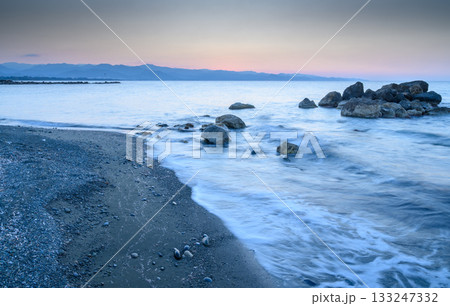 Sunrise paints the coastline with soft hues and tranquil waves meeting rocky shore 133247332