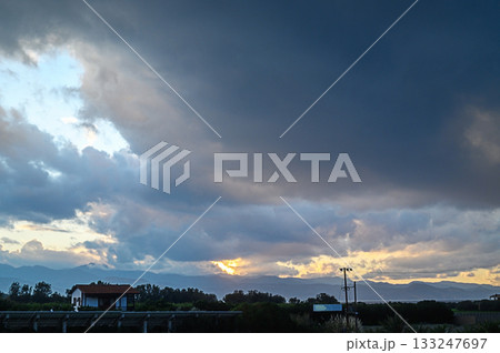 Sunset bathed in vibrant hues behind dark clouds over a rural landscape Sunset bathed in vibrant hues behind dark clouds over a rural landscape 133247697