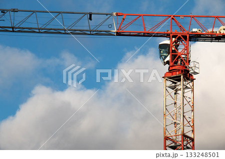 Orthogonal view of Control Cabin of Red-and-white Tower Crane on Construction Site against Blue Cloudy Sky on Sunny Day. Fasteners of Crane and Metal Parts are covered with Rust. 133248501
