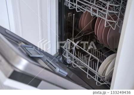 Open dishwasher with clean dishes and utensils arranged in racks in a modern Open dishwasher with clean dishes and utensils arranged in racks in a modern 133249436