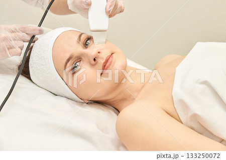 Young caucasian female receiving facial treatment in spa with skincare device 133250072